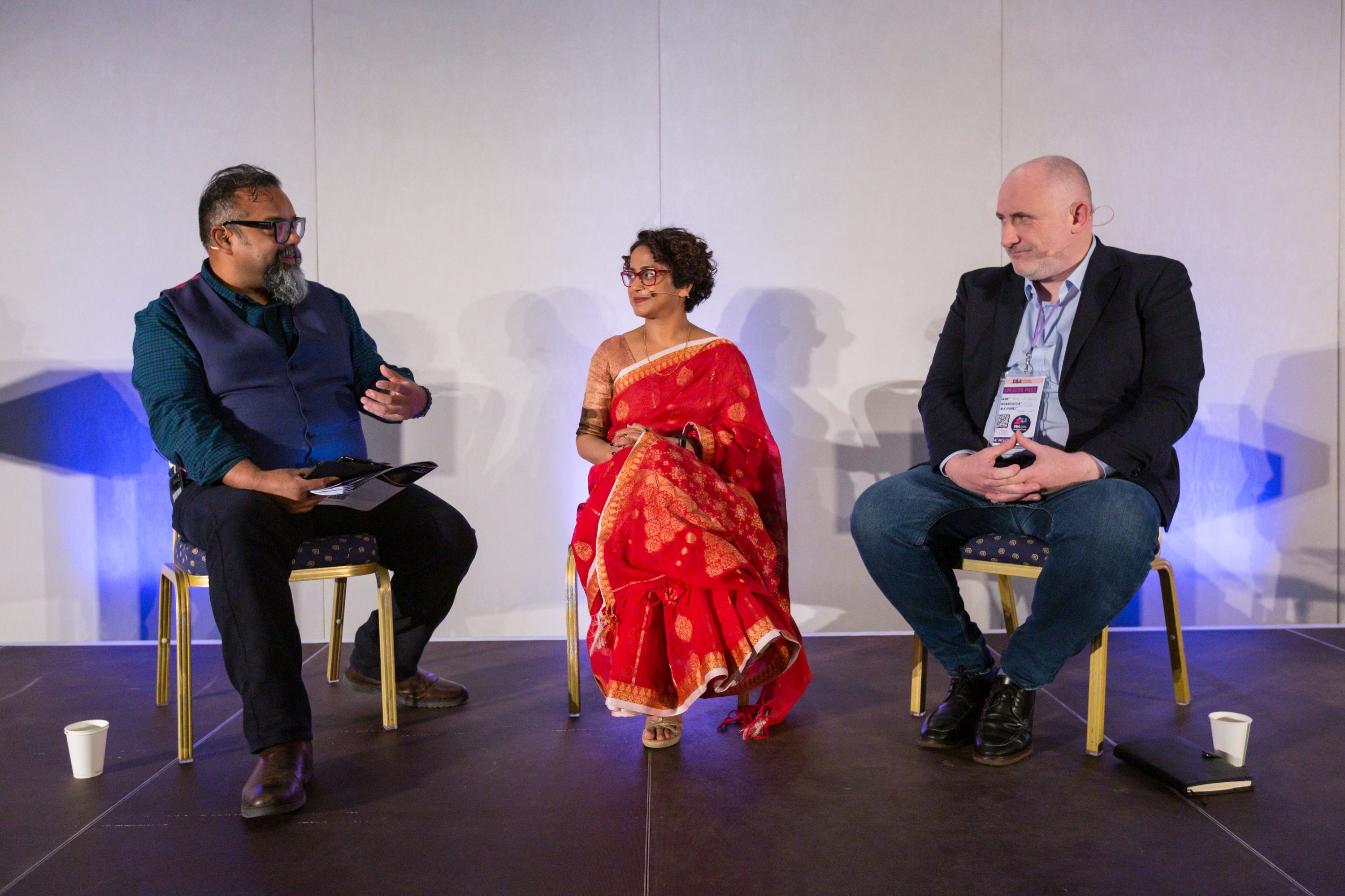 Three people sit on a stage, deep in conversation. They are, from  left to right: Atif, a brown man with dark hair and a grey beard, Poppy, a brown woman with short curly hair wearing a red sari, and David, a white man wearing a suit jacket and jeans. Atif is gesturing as he speaks.