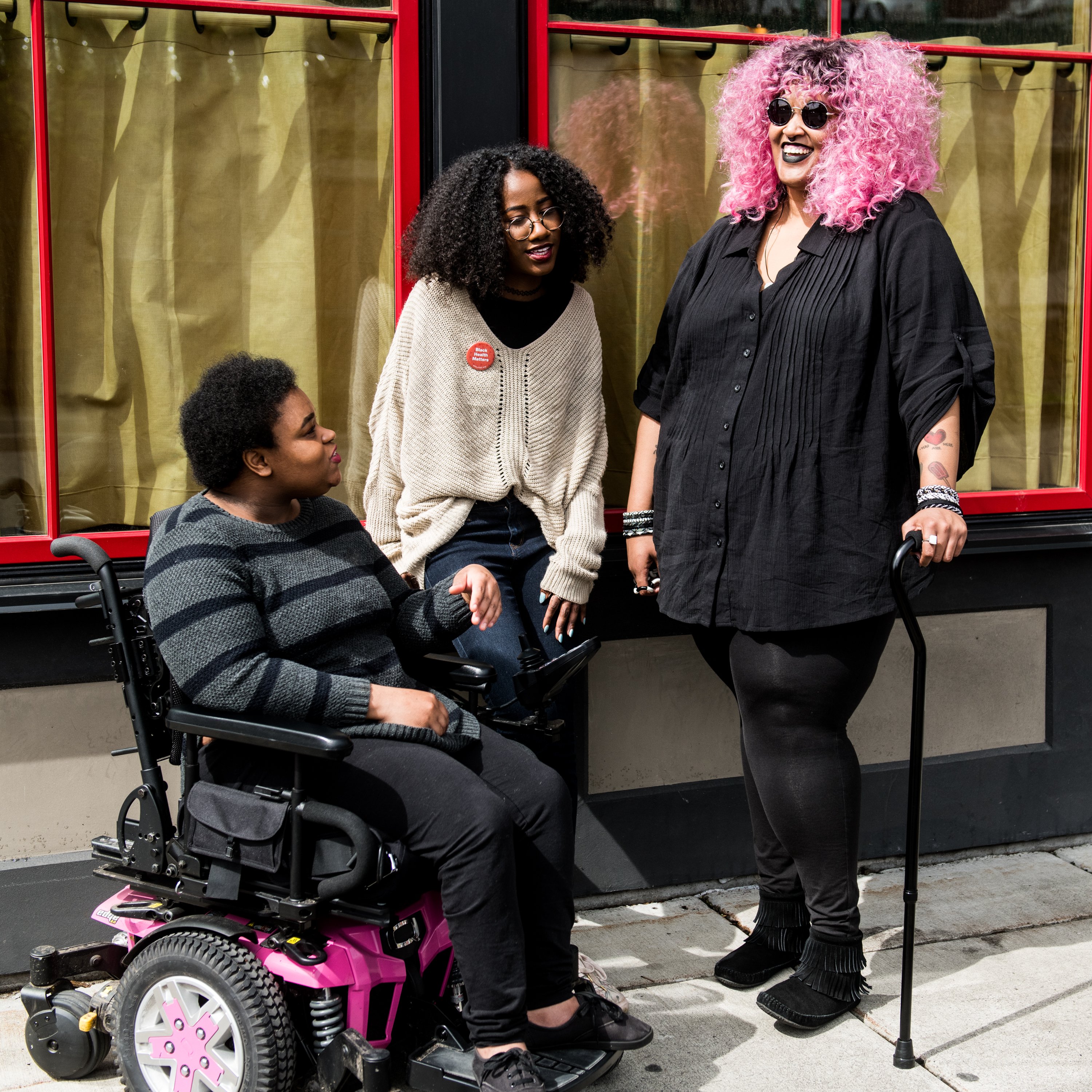Three Black and disabled folx (a non-binary person in a power wheelchair, a femme leaning against a wall, and a non-binary person standing with a cane) engaged in converation. All three are outdoors and in front of a building with two large windows.